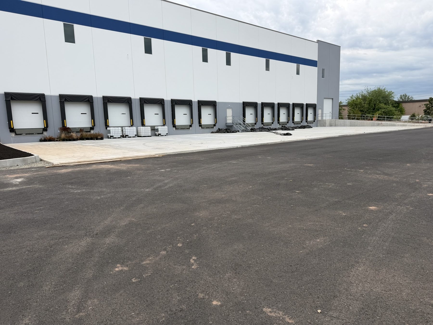 Long precast warehouse elevation with eleven loading dock positions, a drive-in door, new asphalt, and site work in progress.