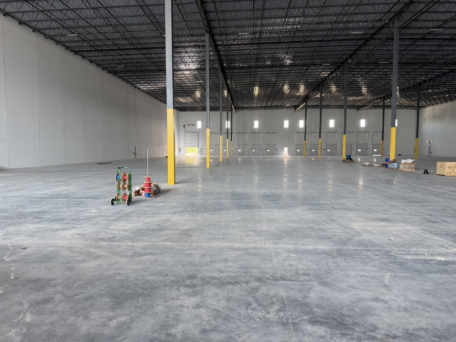 Open warehouse interior with steel columns, open-web roof joists, loading doors, clerestory windows, and a concrete slab.